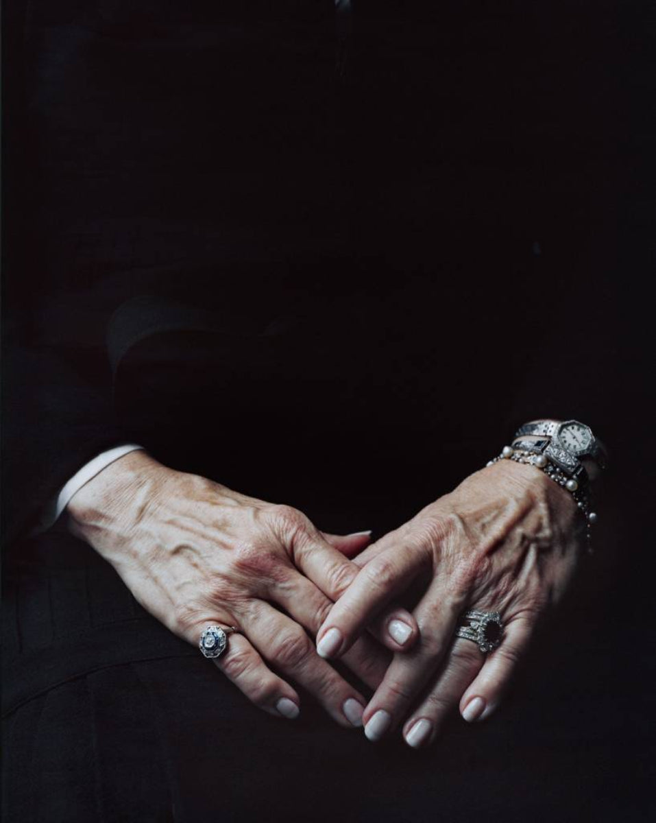 two hands adorned by jewellery resting on black clothed figures lap