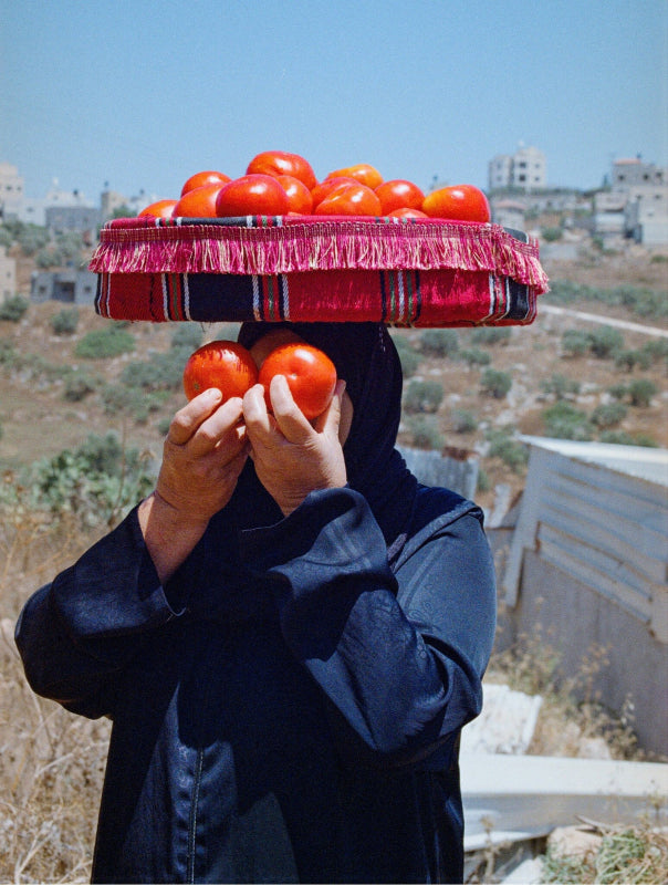 A women holding two oranges in front of her face, carrying a basket of oranges on her head