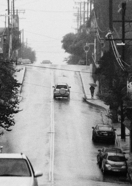 black and white street view with an uphill road with cars and a person with an umbrella