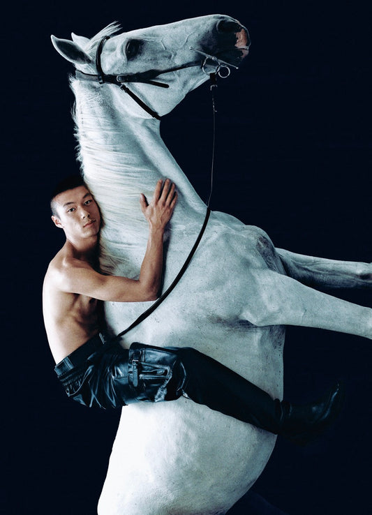 Standing photo with a black background. White horse with a man sitting on top, shirtless wearing black pants.