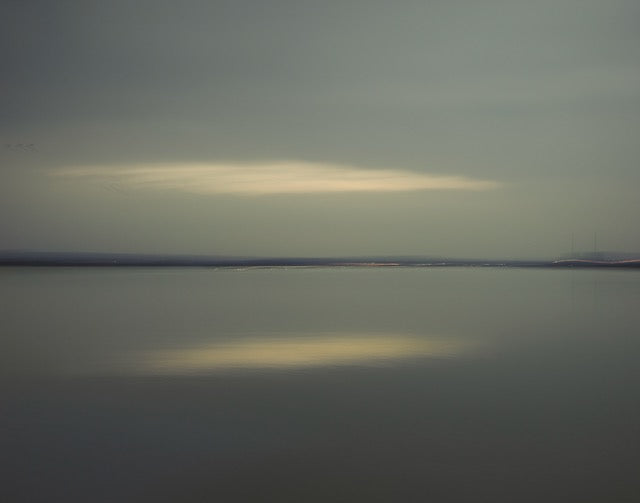 Colour photo of water and a cloudy sky with the horizon in the middle and the reflection in the water of the sun through the clouds.