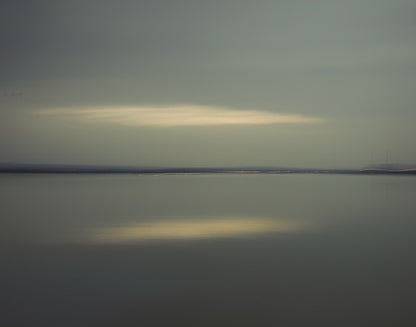 Colour photo of water and a cloudy sky with the horizon in the middle and the reflection in the water of the sun through the clouds.