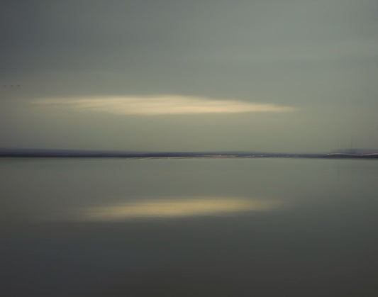 Colour photo of water and a cloudy sky with the horizon in the middle and the reflection in the water of the sun through the clouds.