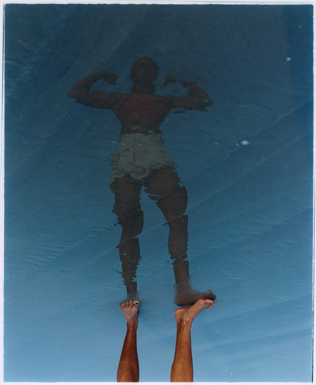 Standing colour image of a reflection of a bodybuilder on a blue ground, wearing white shorts. You see the feet and the rest is reflection.