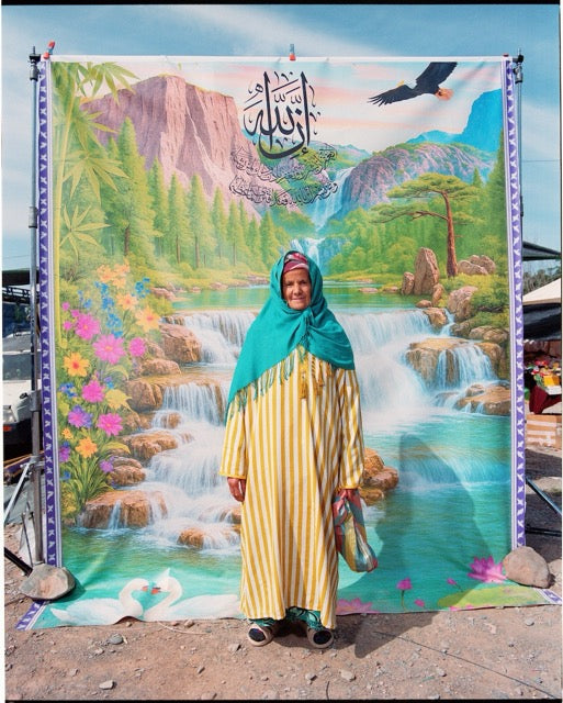 A woman with green scarf and yellow striped robe standing in front of a backdrop of a mountain landscape.