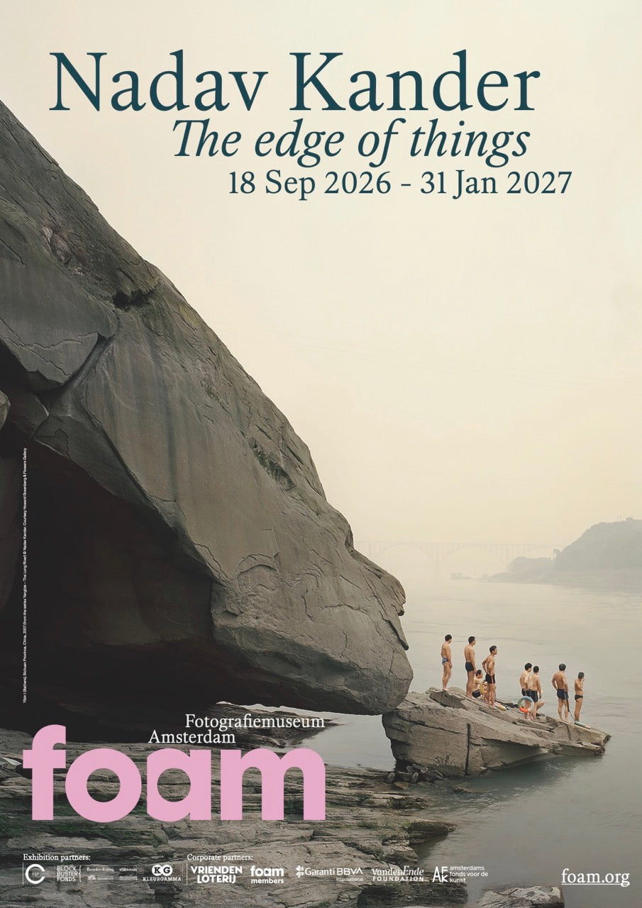 Standing poster of the Nadav Kander exhibition, showing a large grey rock on the side of a river, with a smaller rock in the water. On top of this small rock is a group of 8 people in bathing suits. Dark blue text on the top and pink Foam logo on the bottom, over the image. 