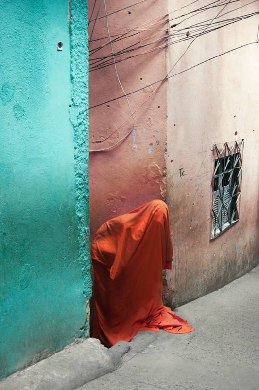 A figure covered in orange fabric in between a pink and blue building