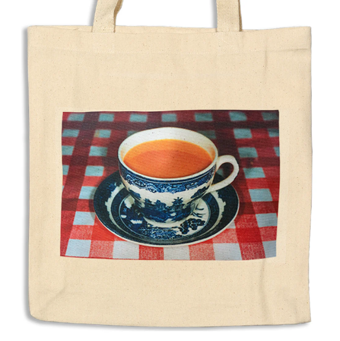 Ecru tote bag with the image of porcelain tea cup and saucer on a red checkered table cloth.